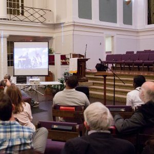 People sitting in pews and watching a slideshow about social justice at a church-hosted event.