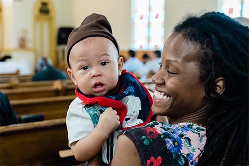 Woman smiling and holding up her baby, who's staring sleepily into the camera, at a church-hosted event.
